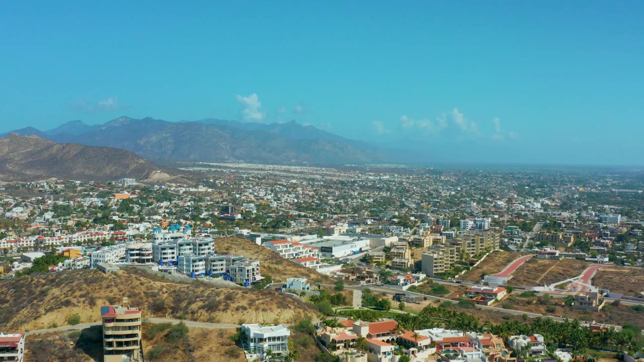 Aerial panorama over Los Cabos, Mexico, with a rounded dry hill in the foreground, dense low-rise buildings, mountain ridges, and no visible people.