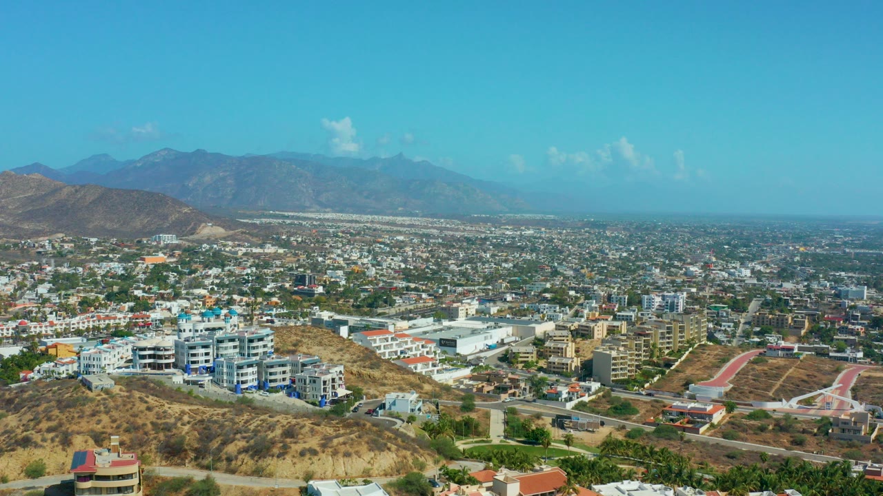 Wide elevated view over Los Cabos, Mexico, with dry hillside foreground, low-rise buildings, distant mountains, blue sky, and no visible people.
