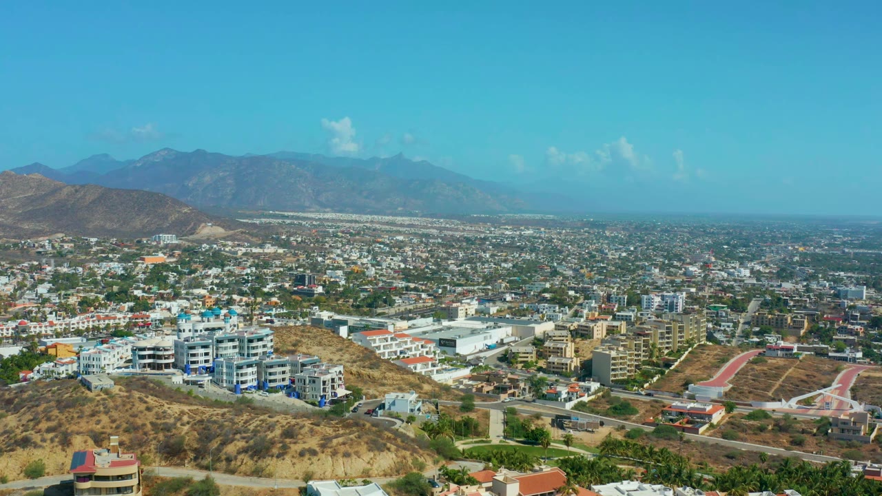 Los Cabos hillside townscape with tan scrubland in front, white apartment-like buildings, scattered streets, hazy mountains, and no visible people.