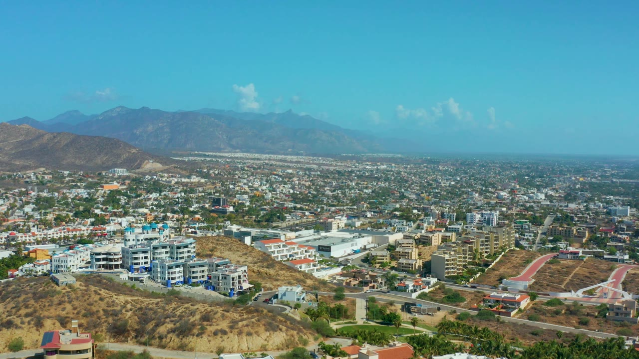 Elevated Los Cabos view showing a broad spread of buildings, a dry hill in the foreground, mountain ridges on the horizon, and no visible people.