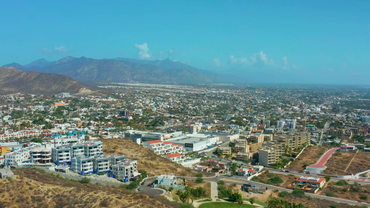 Second frame holds on the Los Cabos city panorama, with white and tan buildings across the midground, arid hills at the edges, mountains beyond, and no visible people.