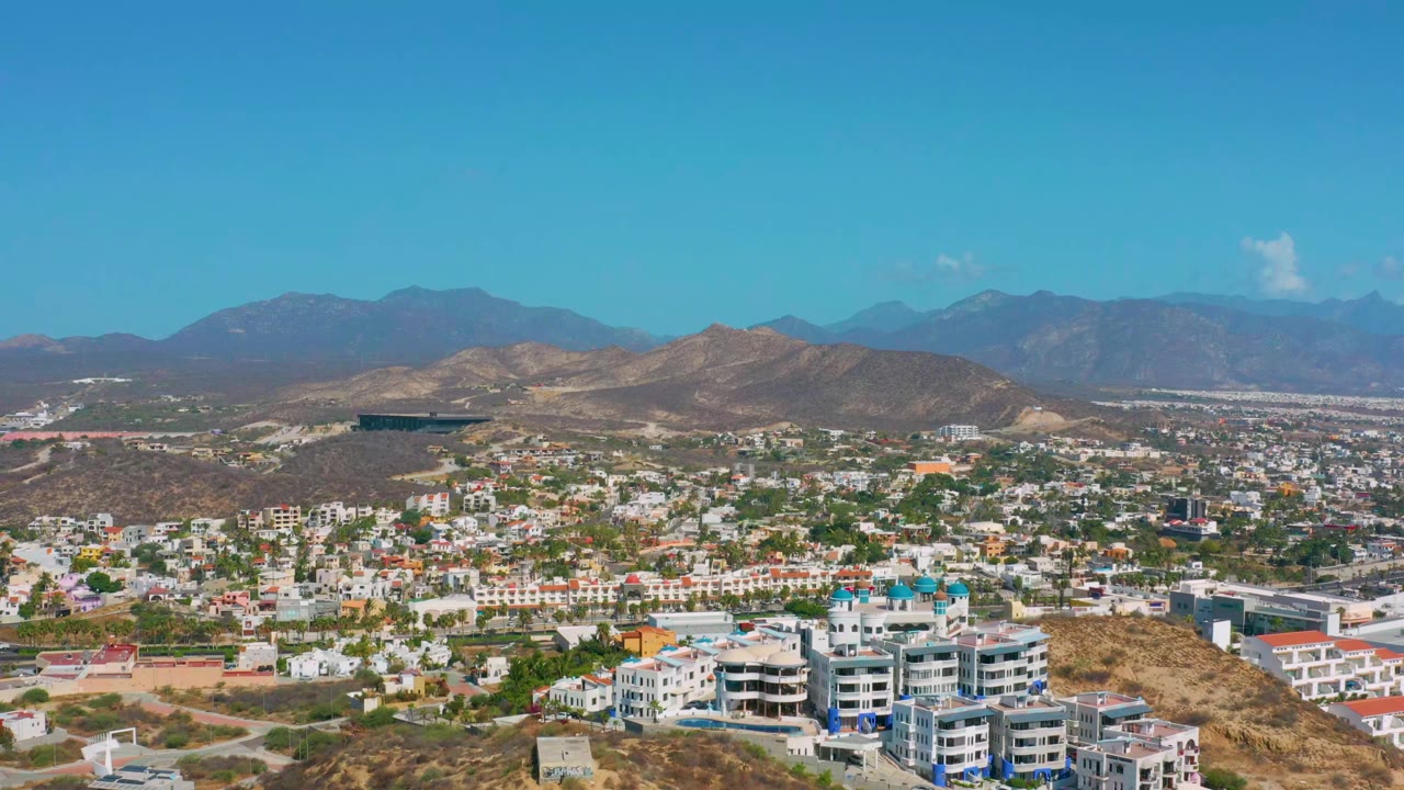 Opening frame of Los Cabos, Mexico, showing modern hillside buildings in the foreground, a spread-out town beyond them, dry hills, blue mountains, clear sky, and no visible people.