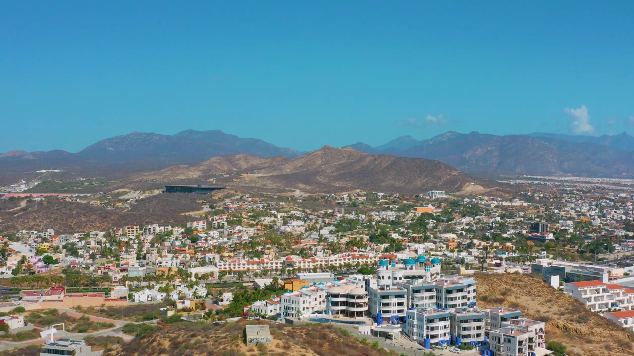 At 0:02, Los Cabos appears in a wide elevated composition with modern buildings along the lower frame, a town grid through the middle, tan hills, blue mountains, clear sky, and no visible people.