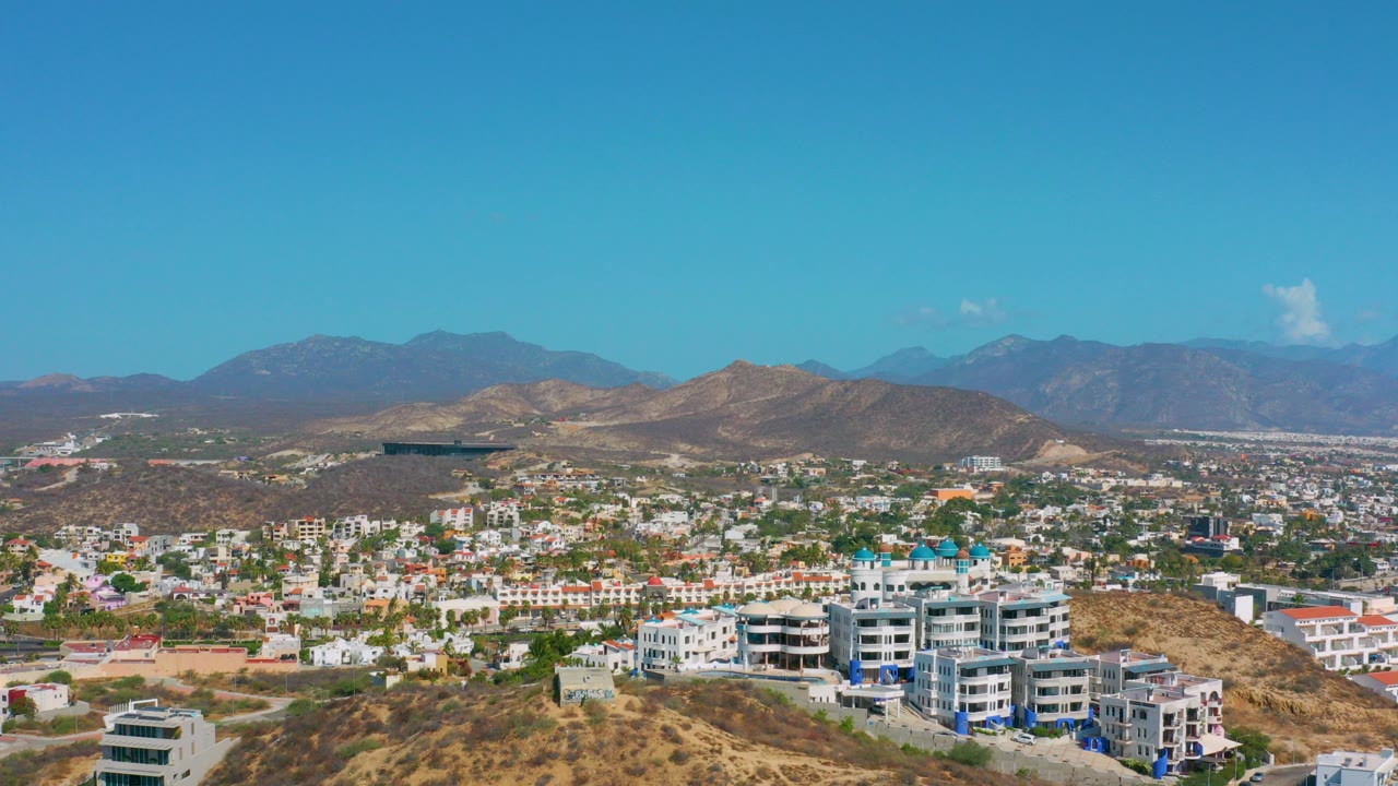 Opening aerial frame over Los Cabos, Mexico, showing low-rise city blocks below dry hills and distant blue mountains under a clear sky, with no visible people.