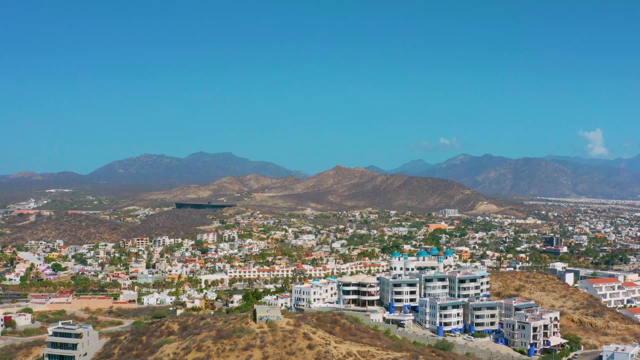 Early aerial view in Los Cabos, Mexico, with white and light-colored buildings across the lower frame, brown hills behind them, and no visible people.