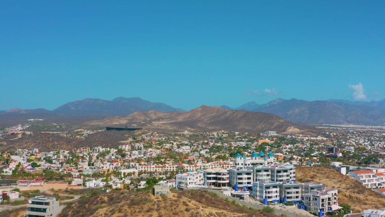 Aerial frame over Los Cabos, Mexico, showing dense low-rise development, scrub-covered ridges, hazy mountains, and a broad clear sky with no visible people.