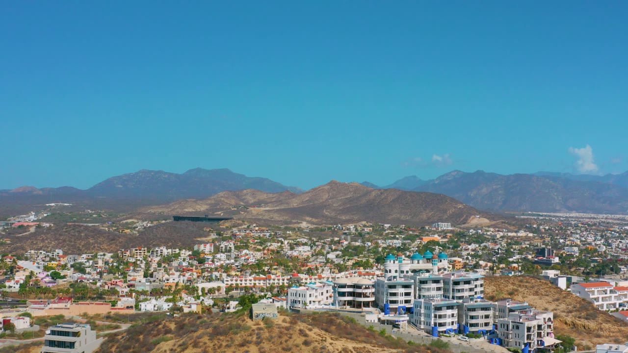 Aerial panorama of city blocks, hillside buildings, mountain backdrop, and bright blue sky.