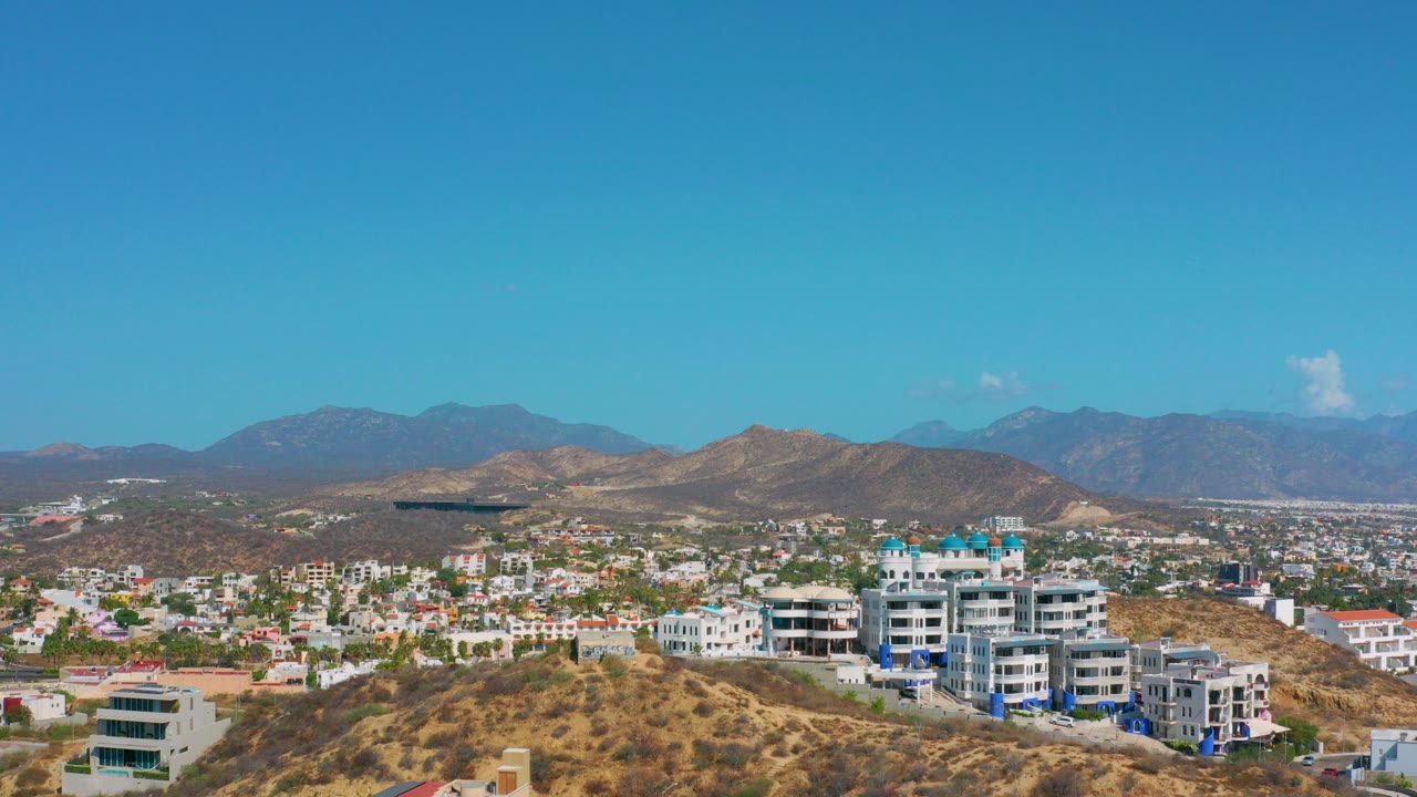 Opening frame of Los Cabos, Mexico showing hillside homes and modern buildings beneath a clear blue sky, with distant mountains and no visible people.
