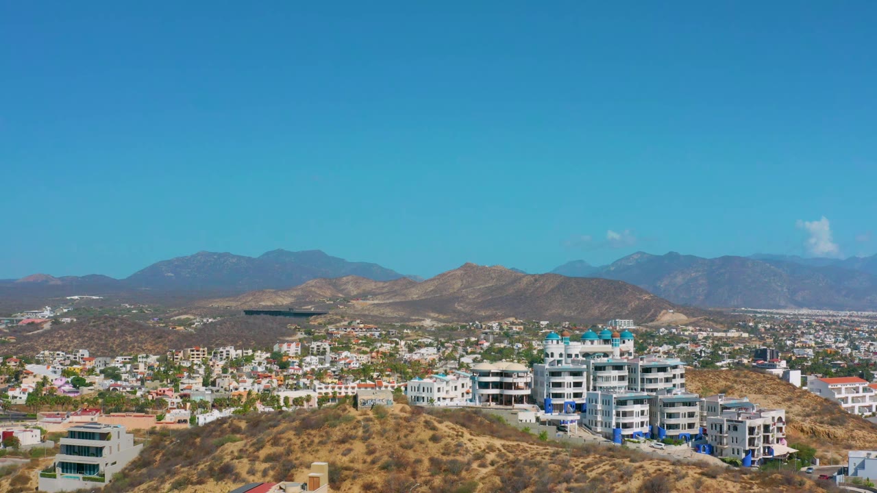 Wide aerial view of white buildings on dry hills with mountains in the background.