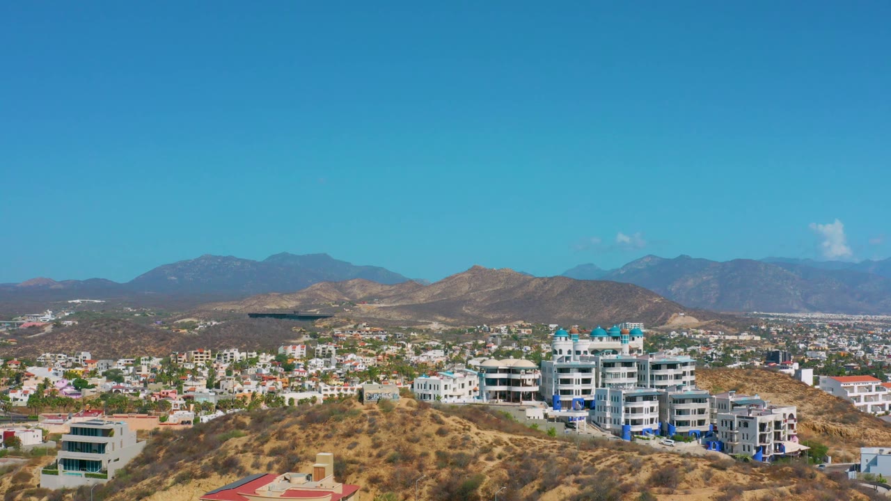 A modern multi-story complex stands on the Los Cabos hillside, backed by scattered city blocks, dry ridges, and a cloudless blue sky with no visible people.