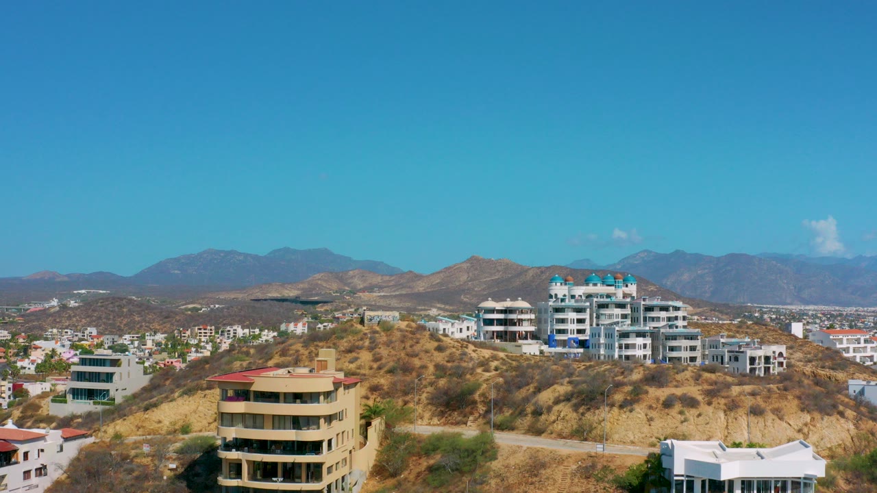Los Cabos hillside view with tan and white residential buildings, dry brush, distant mountains, and no visible people at the start of the clip.