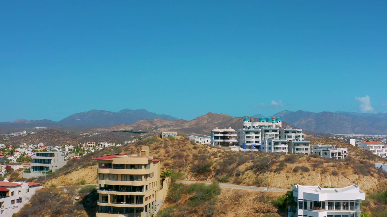 Los Cabos hillside residences fill the lower frame, with white condo buildings on the ridge, dry terrain, far mountains, and no visible people.