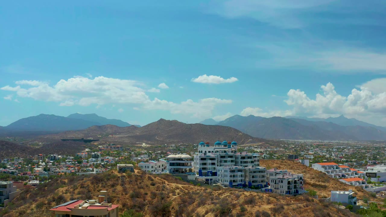 Opening view of Los Cabos, Mexico, showing white resort buildings on a brown hillside, a spread of city blocks, distant blue mountains, bright clouds, and no visible people.