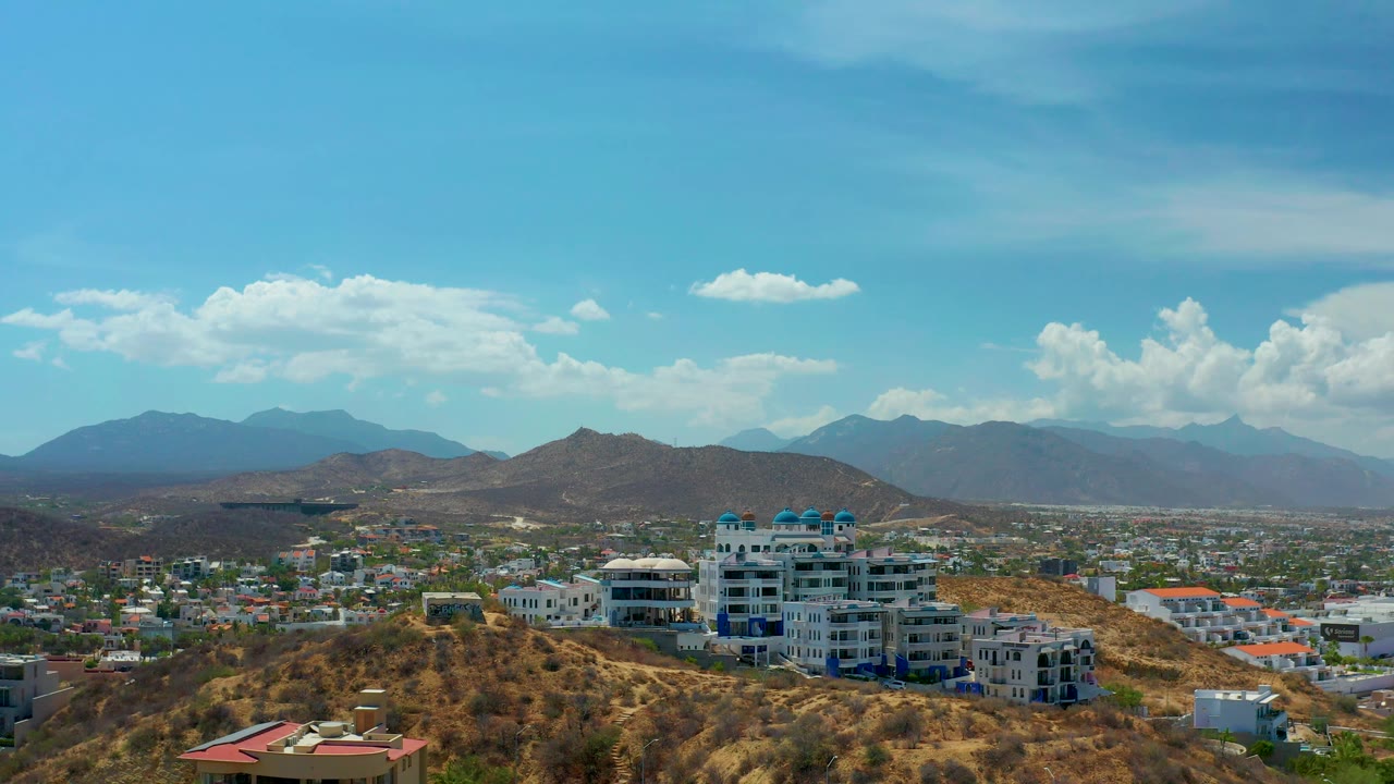 Los Cabos, Mexico, appears from an elevated wide view with white hillside resort structures, low buildings across the valley, brown ridges, distant mountains, and no visible people.
