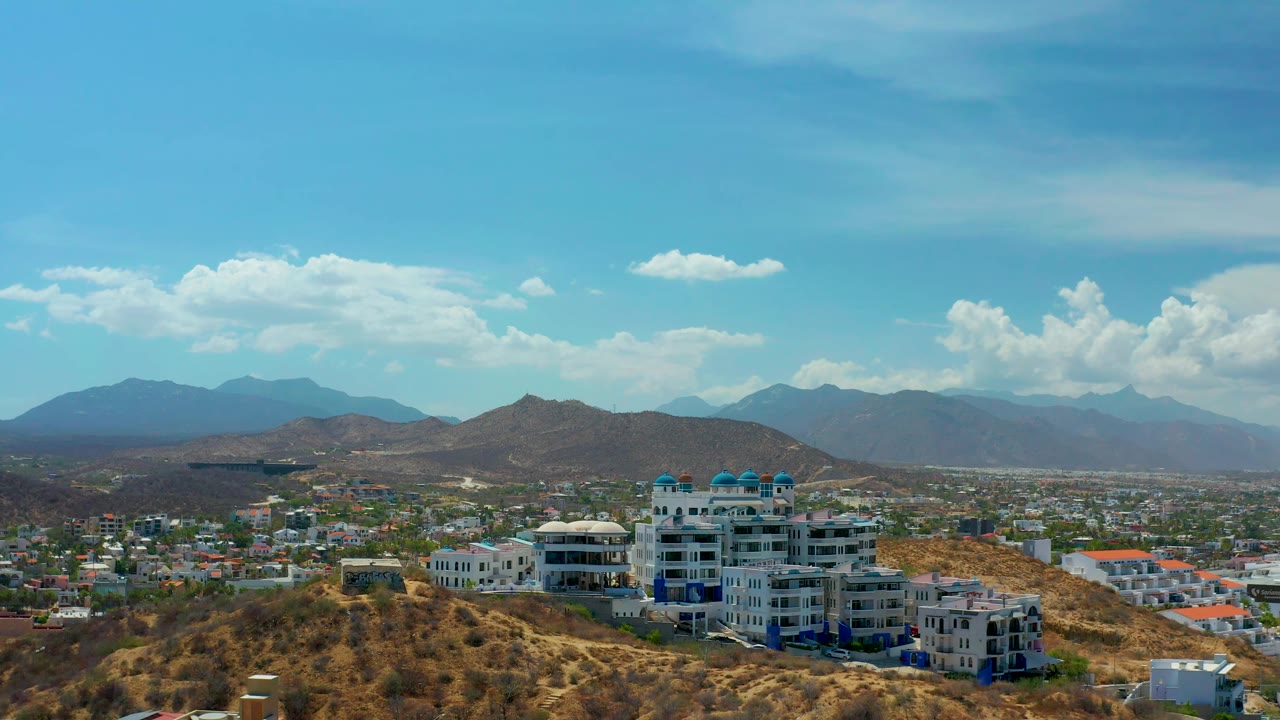 Los Cabos, Mexico fills the frame with hillside residences at lower right, a broad city grid across the middle distance, hazy mountains, scattered clouds, and no visible people.