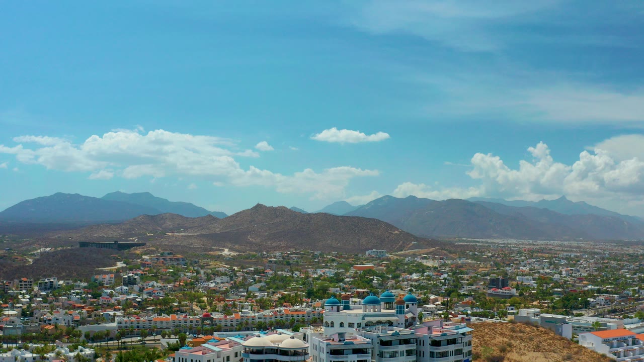 Opening aerial frame over Los Cabos, Mexico, showing low-rise city blocks below dry hills, distant mountains, white clouds, bright blue sky, and no visible people.