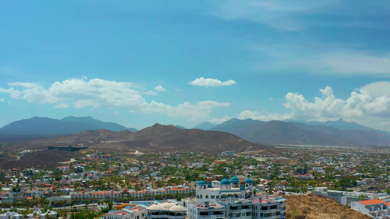 A Los Cabos aerial view one second in, with more of the central hill ridge visible above the city, layered blue mountains in back, and no visible people.