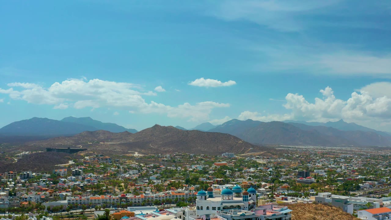 Aerial panorama over Los Cabos, Mexico, with a rounded brown hill centered above the urban grid, mountains fading into haze, white cloud banks, and no visible people.