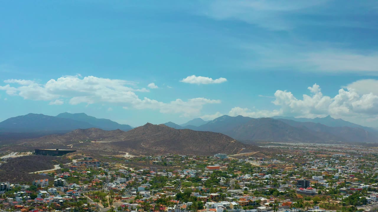Los Cabos, Mexico appears from above with rooftops and roads in the lower frame, rugged brown ridges in the center, hazy mountain layers behind them, and no visible people.