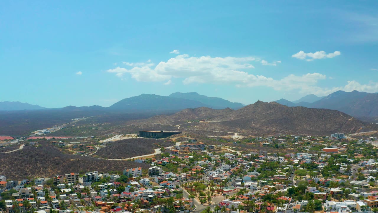 High view over Los Cabos, Mexico, with clustered homes in the lower frame, a rounded brown hill near center, blue mountain ranges in back, and no visible people.