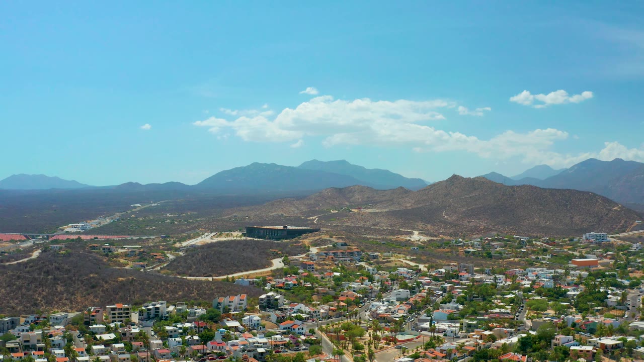 Wide Los Cabos aerial with rooftops and neighborhood streets across the lower frame, rounded arid hills beyond, layered blue mountains in the background, and no visible people.