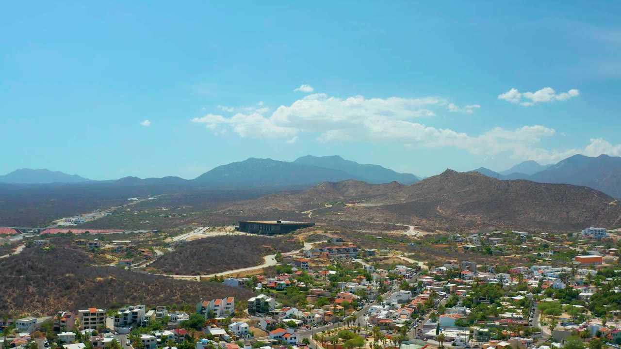 Aerial town edge with winding roads, brown hills, distant ridges, and bright sky.