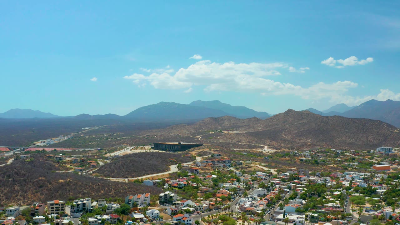 Third frame shows the Los Cabos town spread across the lower frame, a rounded scrub hill in the midground, darker mountain ridges beyond, bright blue sky overhead, and no visible people.