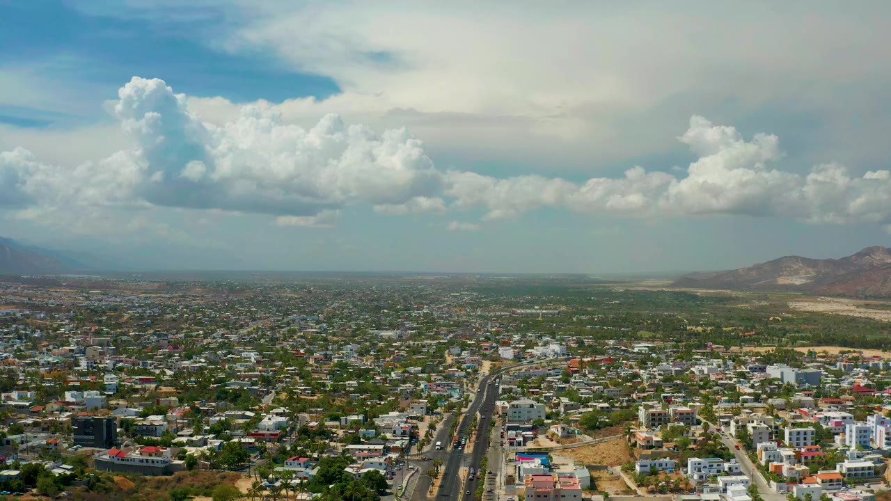 Opening aerial frame over Los Cabos, Mexico, showing a main road, dense low-rise buildings, distant mountains to the right, large clouds, and no visible people.