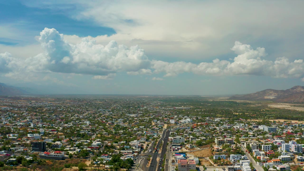Opening aerial frame over a Los Cabos residential district with low buildings, a divided road, distant hills, large clouds, and no visible people.
