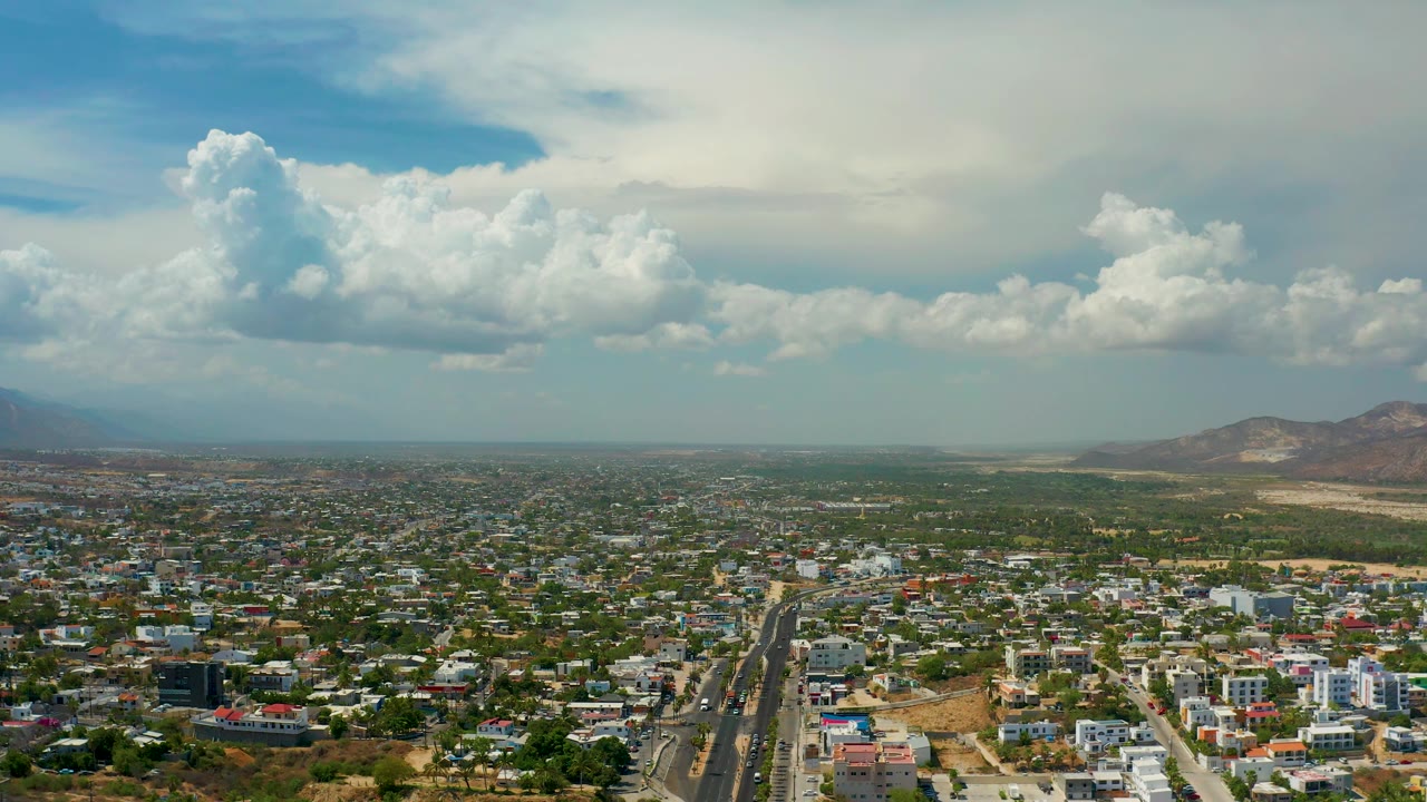 Wide Los Cabos aerial showing dense low-rise residential blocks, dry land beyond town, distant hills, dramatic cloud cover, and no visible people.