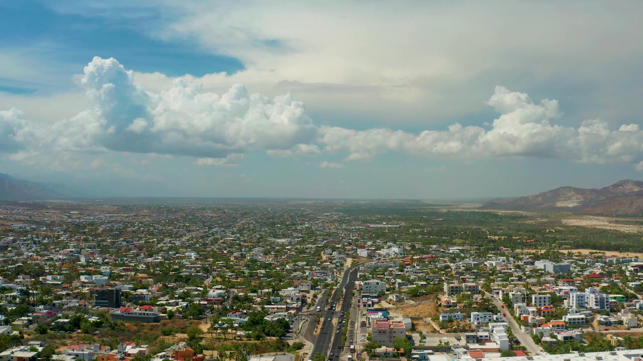 Aerial view over Los Cabos, Mexico, with a dense spread of houses, a straight road in the lower center, low mountains on the right horizon, large white clouds overhead, and no visible people.
