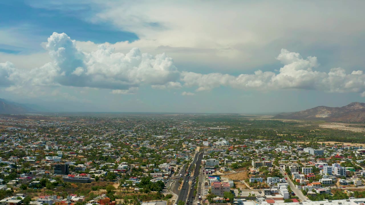 Wide aerial city view with roads, houses, distant mountains, and cloud cover.