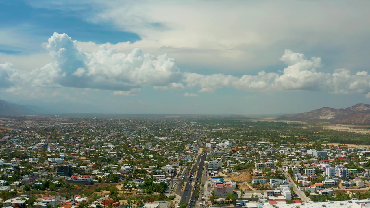 Aerial panorama over Los Cabos, Mexico, with residential blocks across the lower frame, a long straight road near center, hazy distance, mountains to the right, layered white clouds above, and no visible people.