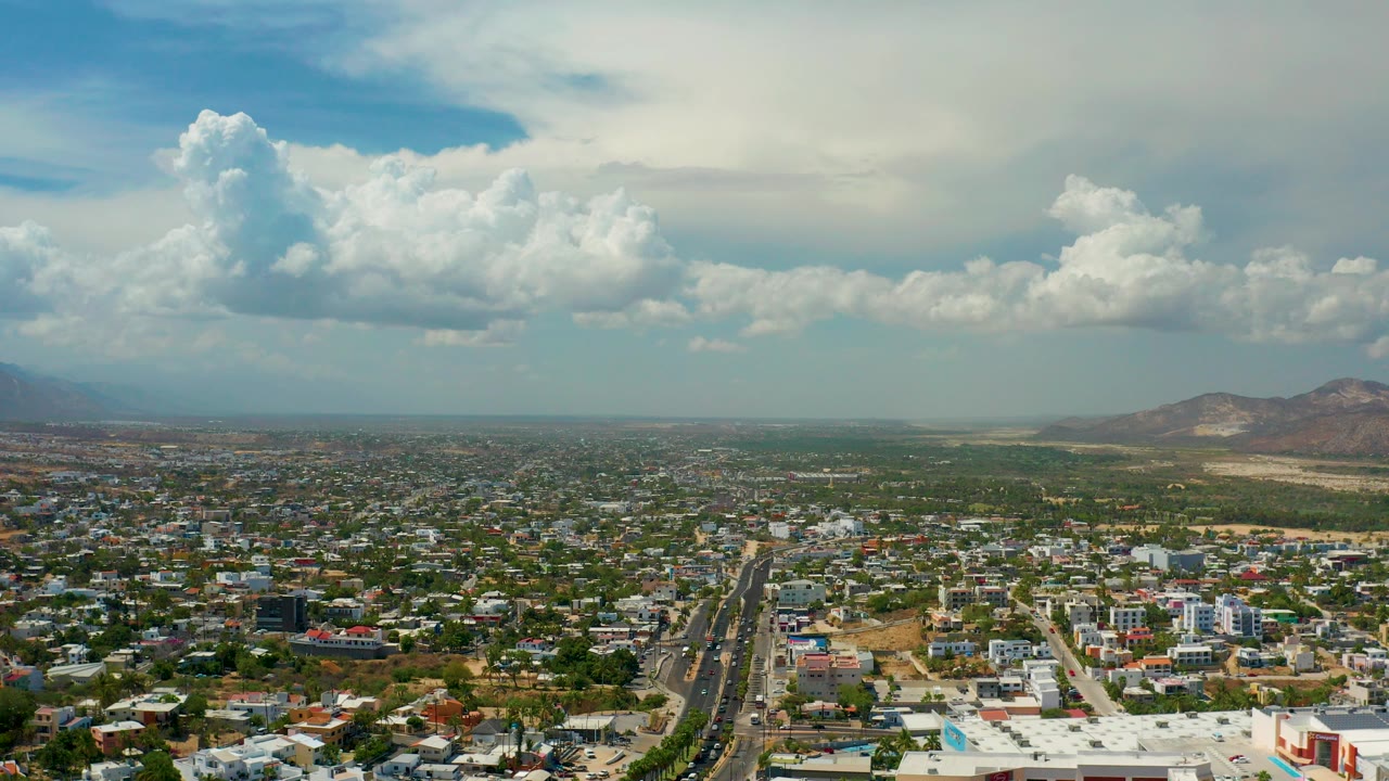 Wide aerial view over Los Cabos, Mexico, with a long divided road through dense buildings, distant hills, large clouds, and no visible people.