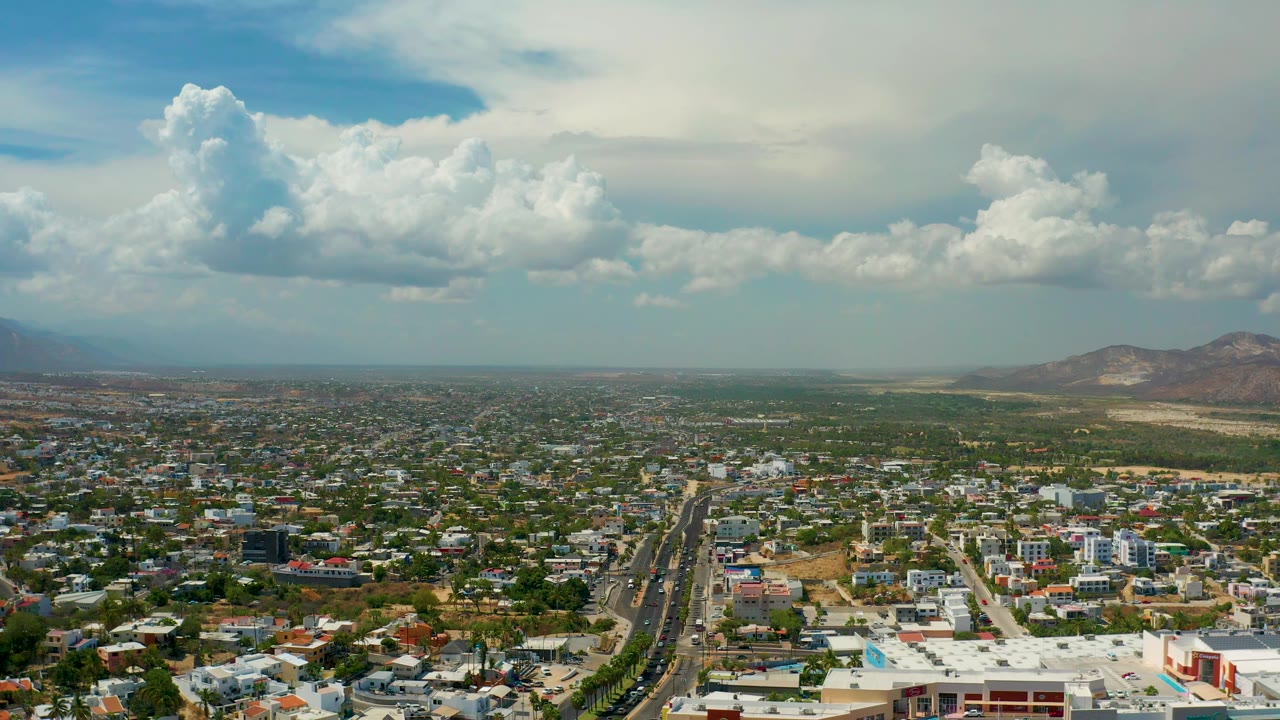 Los Cabos, Mexico appears from above with clustered buildings across the foreground, a broad road at left of center, dry hills on the right horizon, and no visible people.