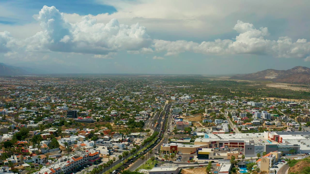 Drone-height city view with a central roadway, rooftops, green patches, mountains, and cloud cover.