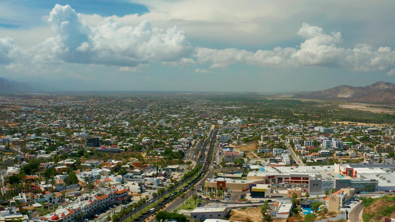 High aerial view of Los Cabos, Mexico, with a broad road cutting through low buildings, reddish dry ground in the foreground, distant hills, and bright cumulus clouds overhead.