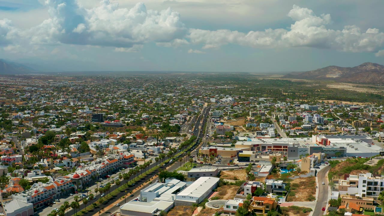 Aerial view of urban blocks, tree-lined road, white roofs, and distant hills.