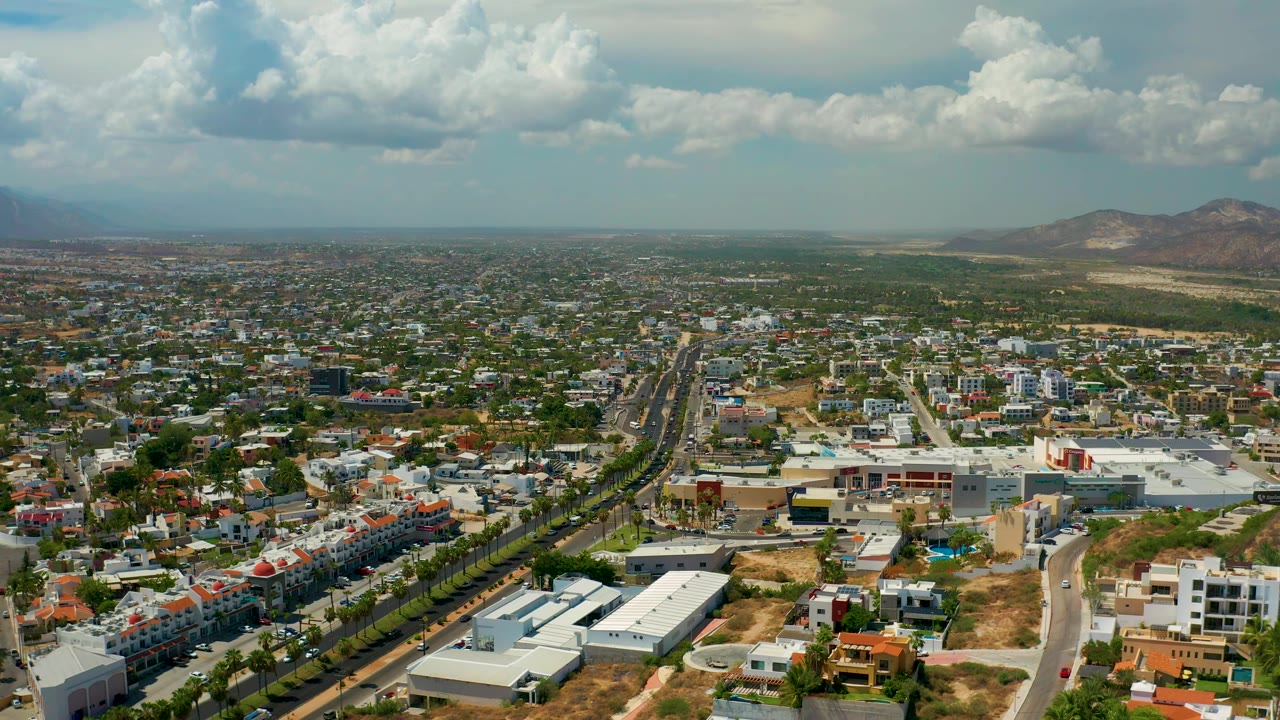 Aerial frame over Los Cabos, Mexico, showing streets, residential and commercial-looking buildings, dry lots, distant hills, and layered clouds, with no visible people.