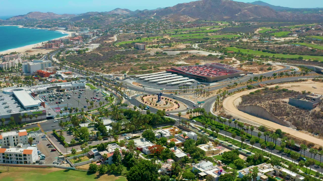 Opening aerial frame over Los Cabos, Mexico, showing palm-lined roads, low buildings, a curved shoreline, green patches, and mountains in the distance with no visible people.