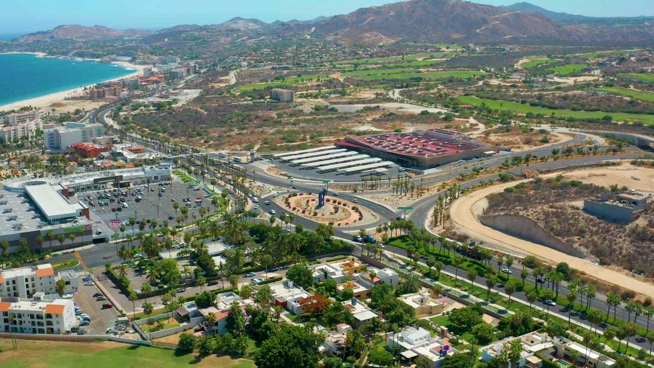 Opening aerial frame over Los Cabos, Mexico, with turquoise water and beach at left, roads and buildings below, green fields inland, mountains on the horizon, and no visible people.