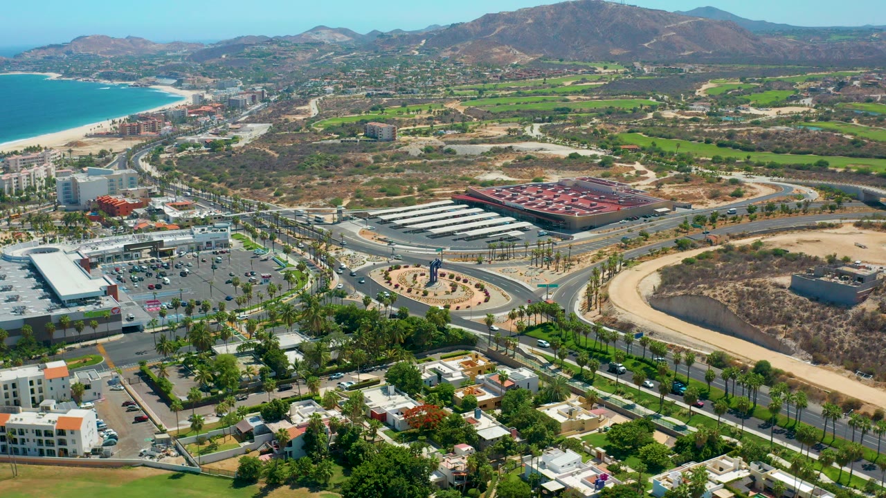 High aerial view over Los Cabos, Mexico, with palm-lined roads, a roundabout, commercial buildings, beach water at left, mountains in the distance, and no visible people.