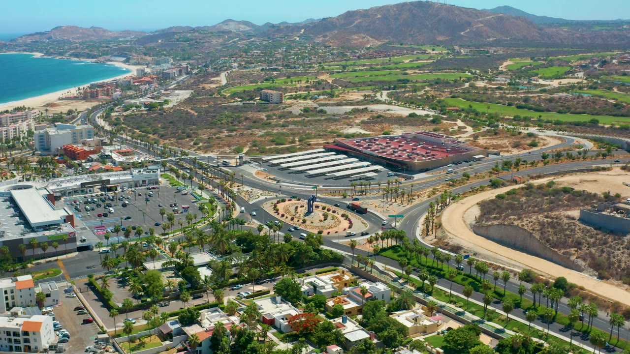 Drone-style view over Los Cabos, Mexico, looking past a road circle and shopping-area buildings toward green fields, brown hills, blue ocean water, and no visible people.