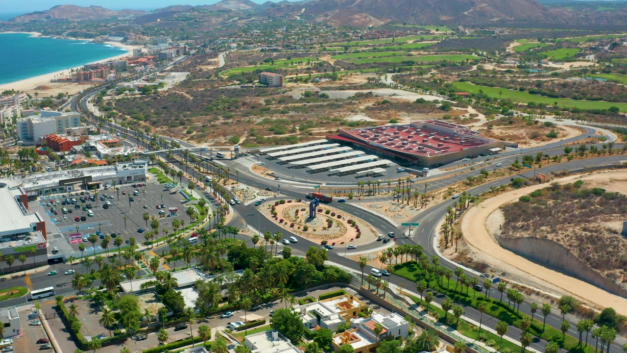 High aerial frame over Los Cabos with a curving road, roundabout, parking lots, commercial buildings, beach at upper left, dry hills beyond, and no visible people.