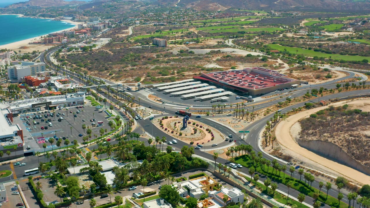 Wide Los Cabos frame showing turquoise water at upper left, roads and traffic through the center, a red-roofed commercial building, parking areas, scrubland, green fields, hills, and no visible people.