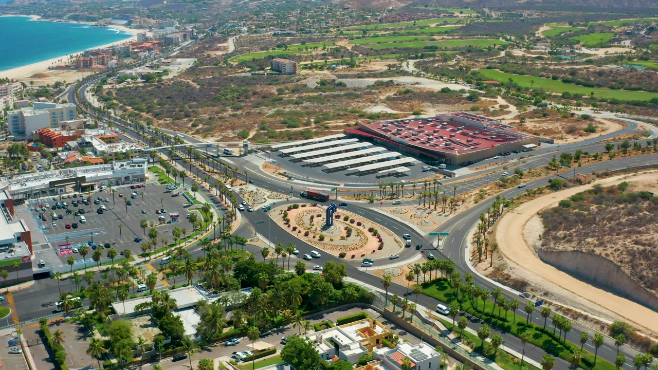 Overhead coastal city view with a roundabout, palm-lined roads, parking, and dry open land.