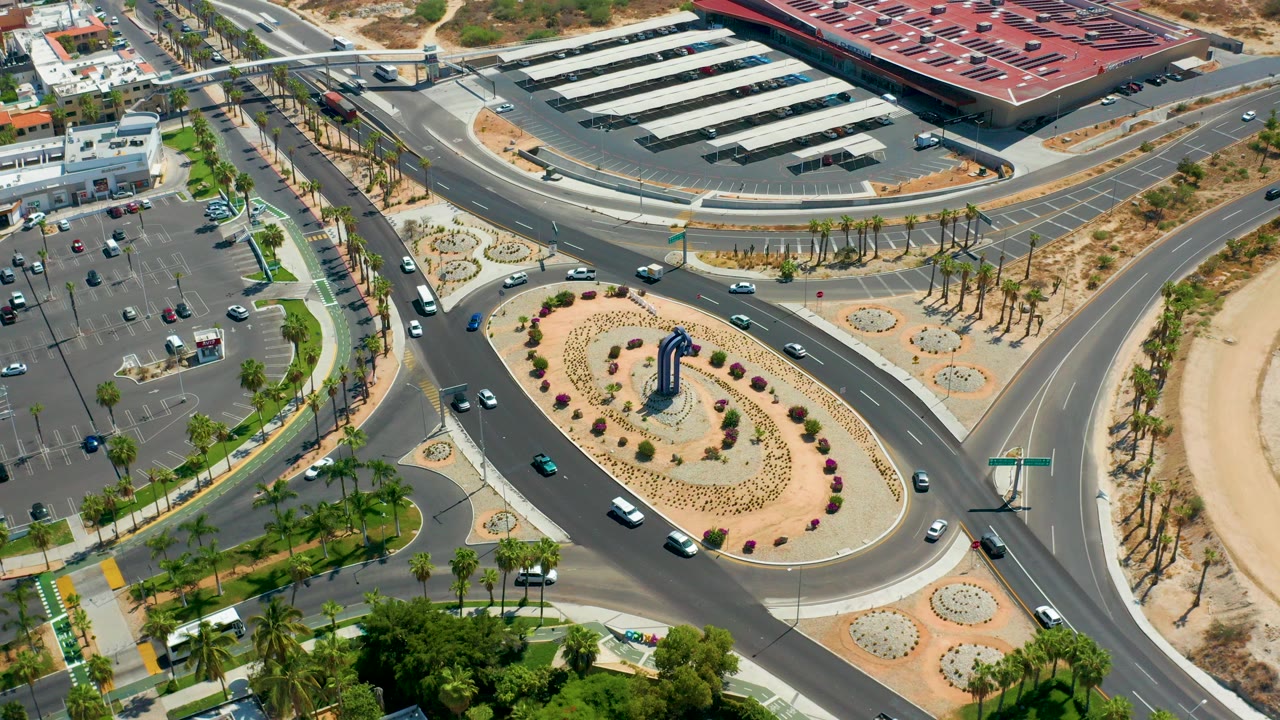 Aerial view in Los Cabos, Mexico showing cars on a curving roundabout bordered by desert planting, palm trees, parking lots, and low buildings, with no visible people.