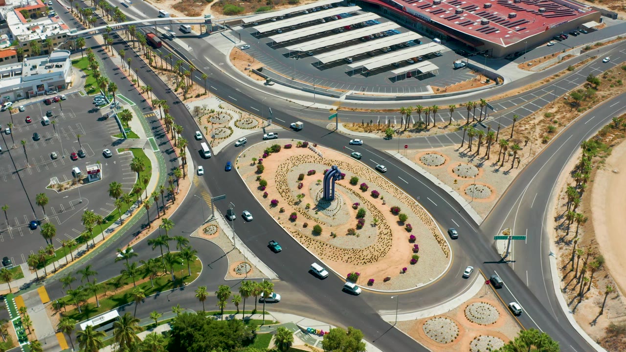 Aerial Los Cabos frame showing vehicles distributed across the roundabout and feeder roads, with a large parking lot, red-roofed building edge, palms, and pale desert landscaping, with no visible people.