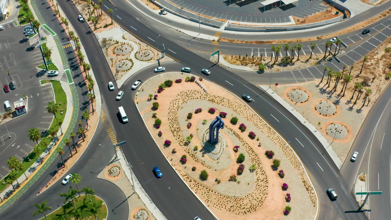 High overhead view in Los Cabos, Mexico, showing cars and vans moving around a landscaped desert median with palm-lined sidewalks and no visible people.