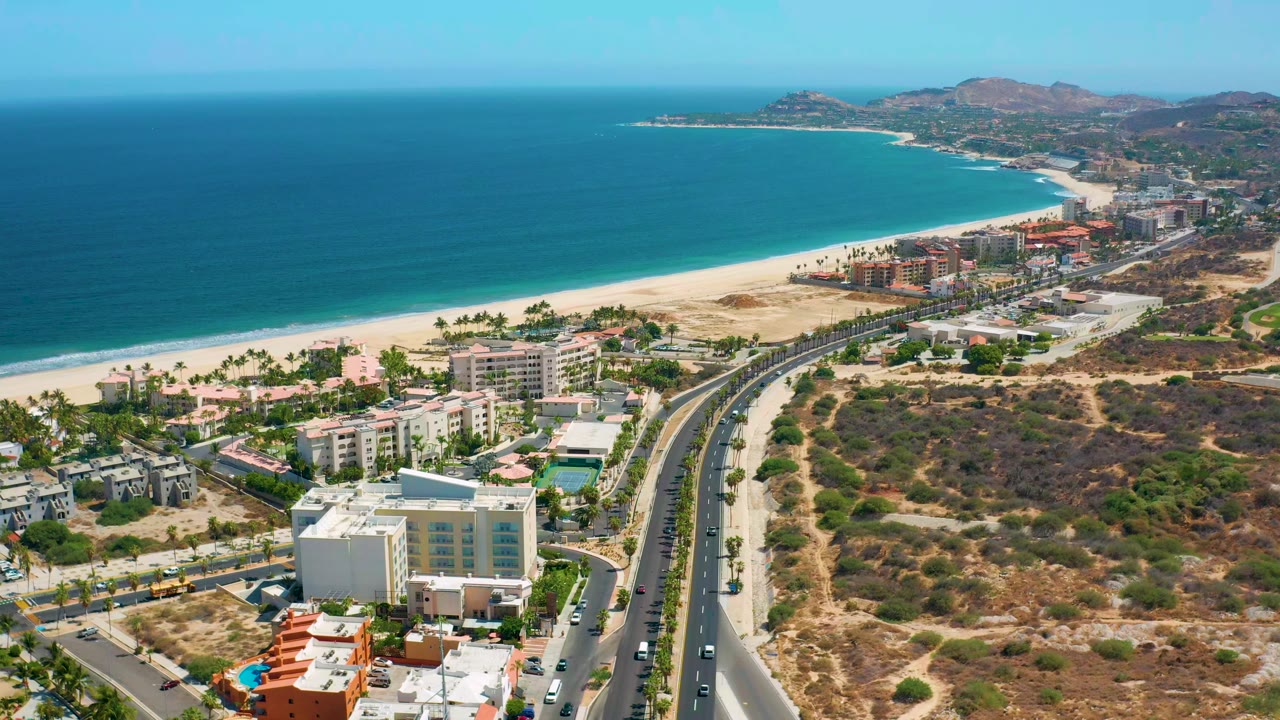 Opening aerial frame over Los Cabos, Mexico, with a divided road in the foreground, low-rise buildings beside it, a long sandy beach, blue sea, distant hills, and no visible people.